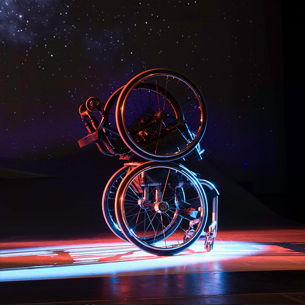 Two silver manual wheelchairs, stacked, make an infinity shape with colorful stage light glinting off their spokes and a sky of constellations sparkling in the background. Photo by BRITT / Jay Newman.